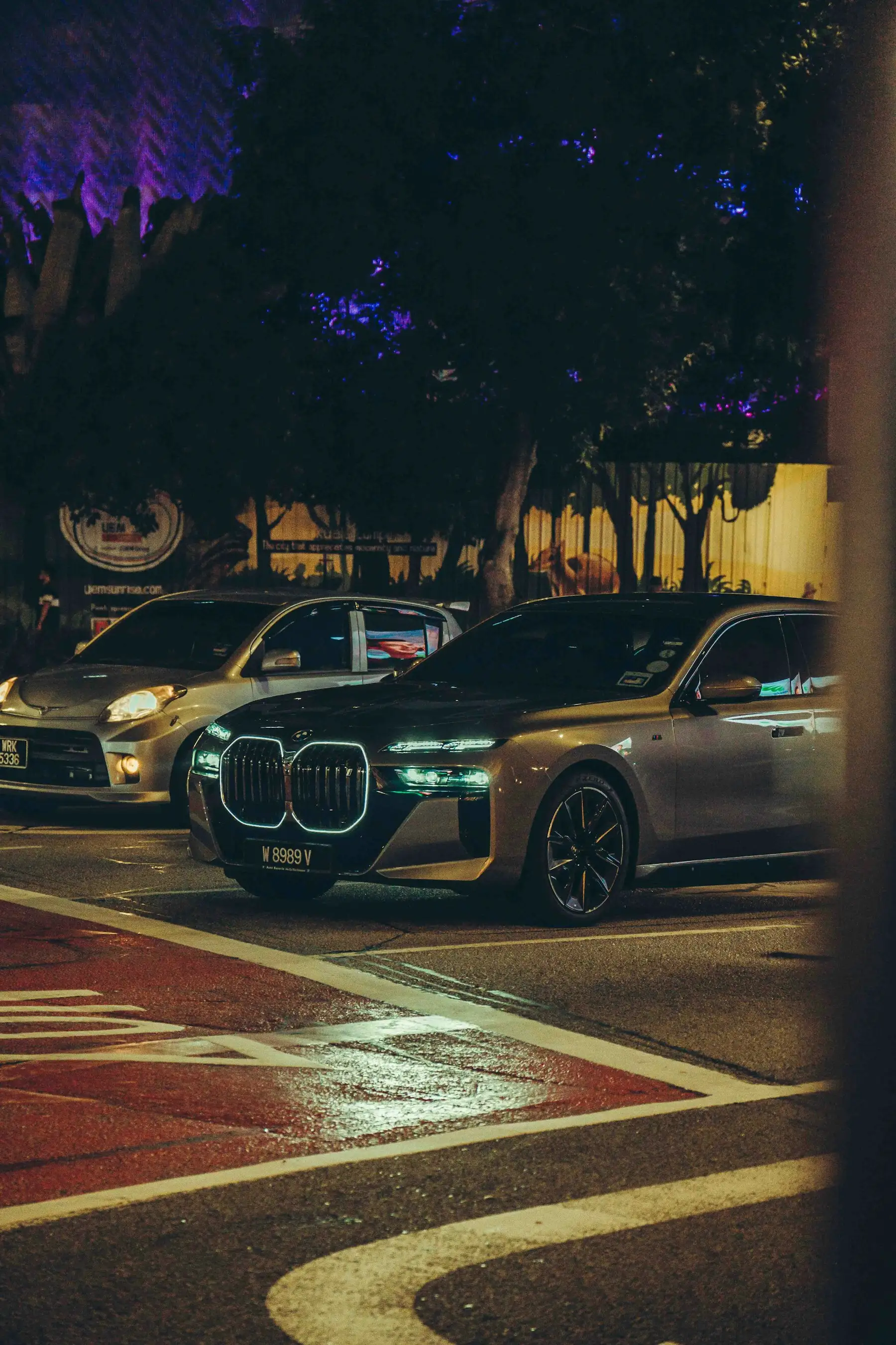 Carro estacionado em avenida urbana à noite, com reflexos das luzes da cidade no asfalto molhado